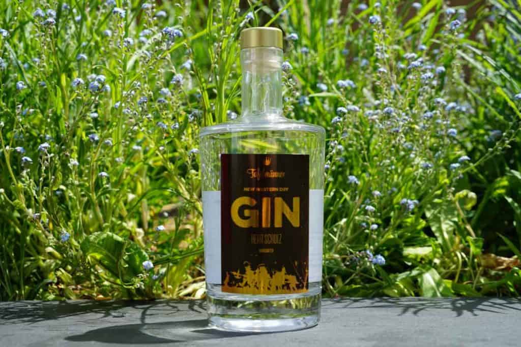 Eine durchsichtige Gin-Flasche mit goldenem Verschluss und dunklem Etikett steht im Freien auf einem dunklen Untergrund, umgeben von hohem grünem Gras und kleinen blauen Blumen im Hintergrund.