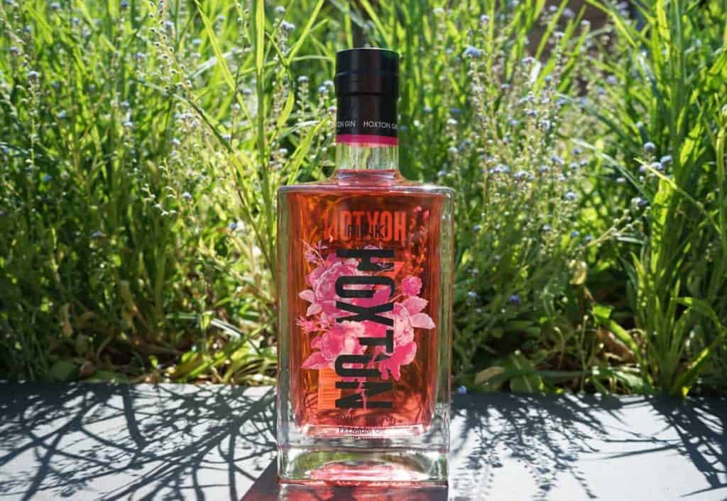 Eine Flasche rosa Hoxton-Gin mit floralen Motiven steht im Freien auf einer dunklen Fläche, umgeben von hohem grünem Gras und Sonnenlicht.