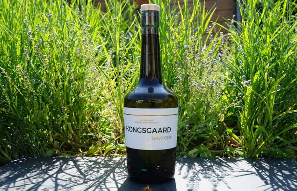 Eine Flasche Kongsgaard Raw Gin steht im Freien auf einer dunklen Fläche, umgeben von hohem grünem Gras und kleinen violetten Blüten im strahlenden Sonnenlicht.