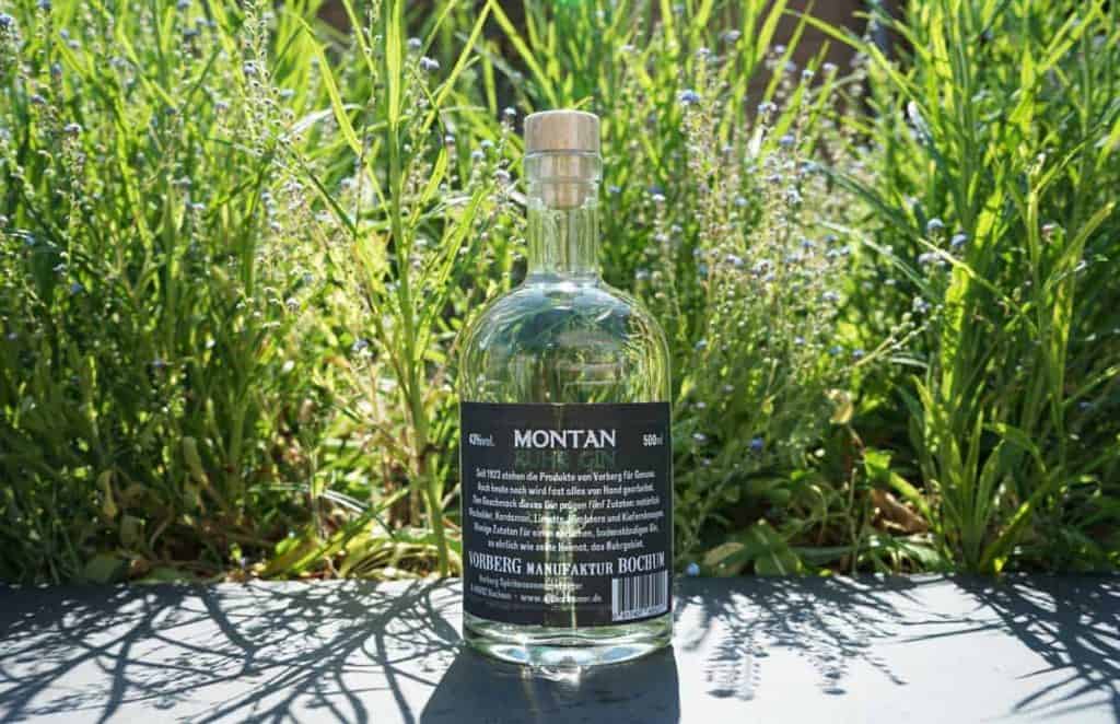 Eine durchsichtige Flasche Montan-Gin mit schwarzem Etikett steht im Freien auf einer dunklen Fläche, umgeben von grünem Gras und Pflanzen im strahlenden Sonnenlicht.
