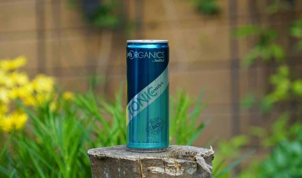 Eine Dose „Organics“-Energy-Drink steht auf einem Baumstumpf, im Hintergrund sind grünes Laub und gelbe Blumen verschwommen zu sehen.