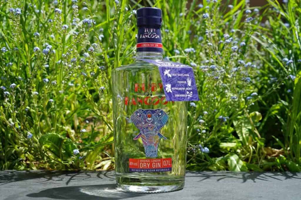 Eine Flasche „Ruby Rangoon“ Dry Gin steht im Freien auf einer Steinfläche, im Hintergrund sind grünes Gras und kleine blaue Blumen zu sehen. Auf der Flasche ist ein Elefant abgebildet, und um den Flaschenhals ist ein blaues Etikett gebunden.