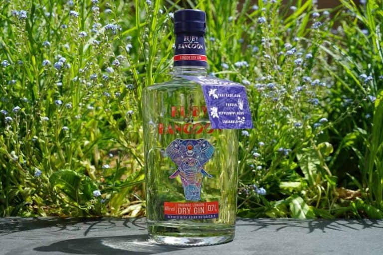 Eine Flasche „Ruby Rangoon“ Dry Gin steht im Freien auf einer Steinfläche, im Hintergrund sind grünes Gras und kleine blaue Blumen zu sehen. Auf der Flasche ist ein Elefant abgebildet, und um den Flaschenhals ist ein blaues Etikett gebunden.