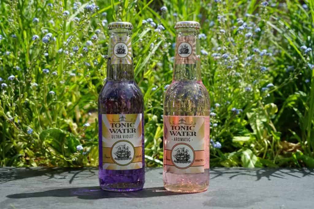 Zwei Glasflaschen mit Tonic Water, eine violette und eine rosa, stehen nebeneinander auf einer Steinfläche im Freien, im Hintergrund sind grünes Gras und kleine blaue Blumen zu sehen.