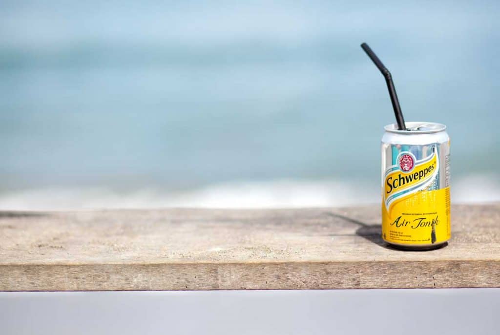 Auf einer Holzfläche steht eine Dose Schweppes mit einem schwarzen Strohhalm, im Hintergrund sind ein verschwommener blauer Ozean und Himmel zu sehen.