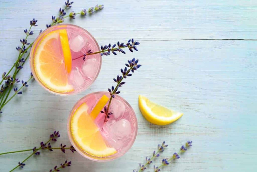 Zwei Gläser rosa Limonade mit Eis, garniert mit Zitronenscheiben und Lavendelzweigen, stehen auf einer hellen Holzunterlage, daneben liegen noch etwas Lavendel und eine Zitronenscheibe.