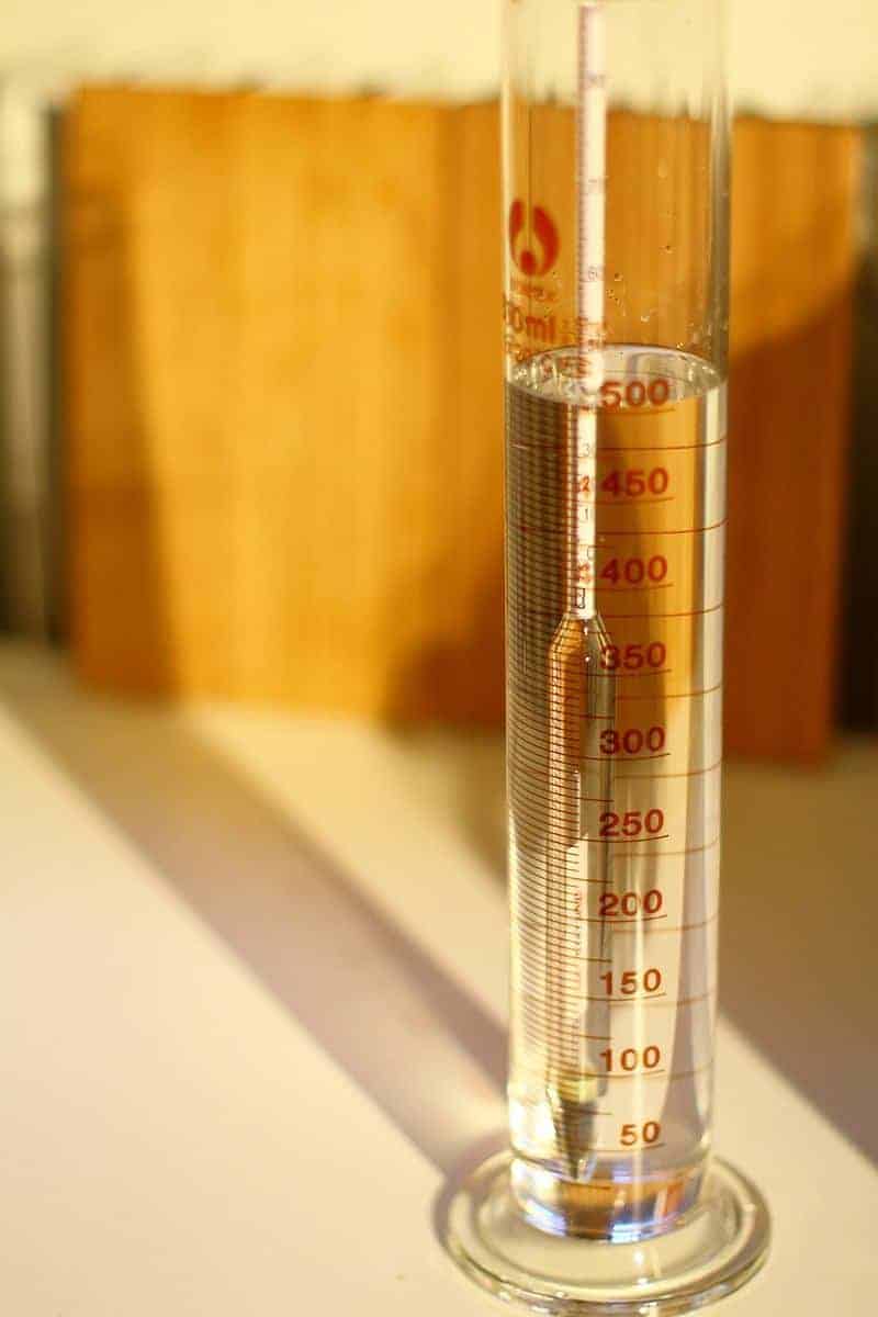 Ein Hydrometer, das in einer klaren Flüssigkeit in einem Messzylinder schwimmt, auf dem rote Messmarkierungen bis 500 ml zu sehen sind. Im Hintergrund sind verschwommene Holzbretter zu erkennen.