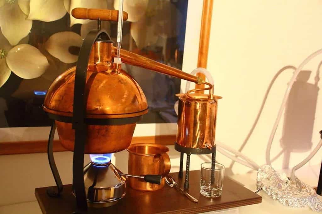 Auf einem Tisch steht eine Kupferbrennblase mit einem Kessel, einem Kondensator und einem Glasgefäß; eine blaue Flamme erhitzt den Kessel. Daneben liegen Tassen, Utensilien und Alufolie; im Hintergrund hängt ein Blumenbild.