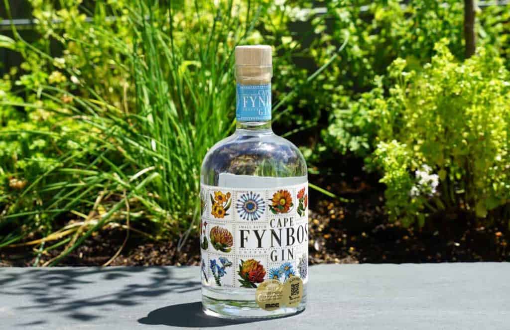 Eine Flasche Cape Fynbos Gin mit einem blumigen Etikett steht im Freien auf einer grauen Fläche, im Hintergrund sind grüne Pflanzen und Kräuter zu sehen.