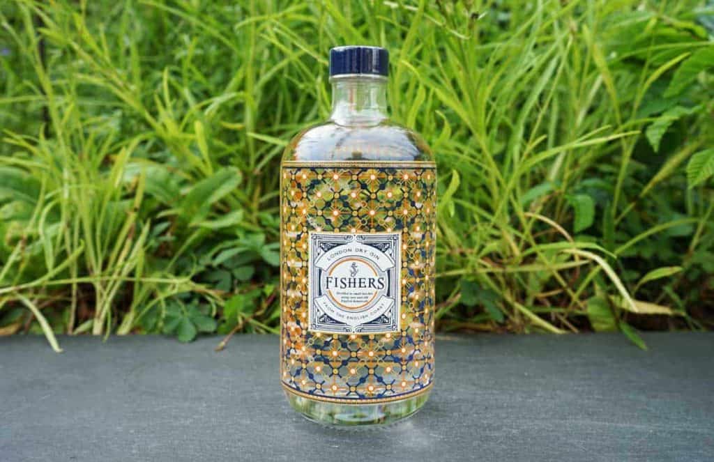 Eine Flasche Fishers Gin mit einem dekorativen Etikett steht im Freien auf einer dunklen Fläche, im Hintergrund sind grünes Gras und Pflanzen zu sehen.