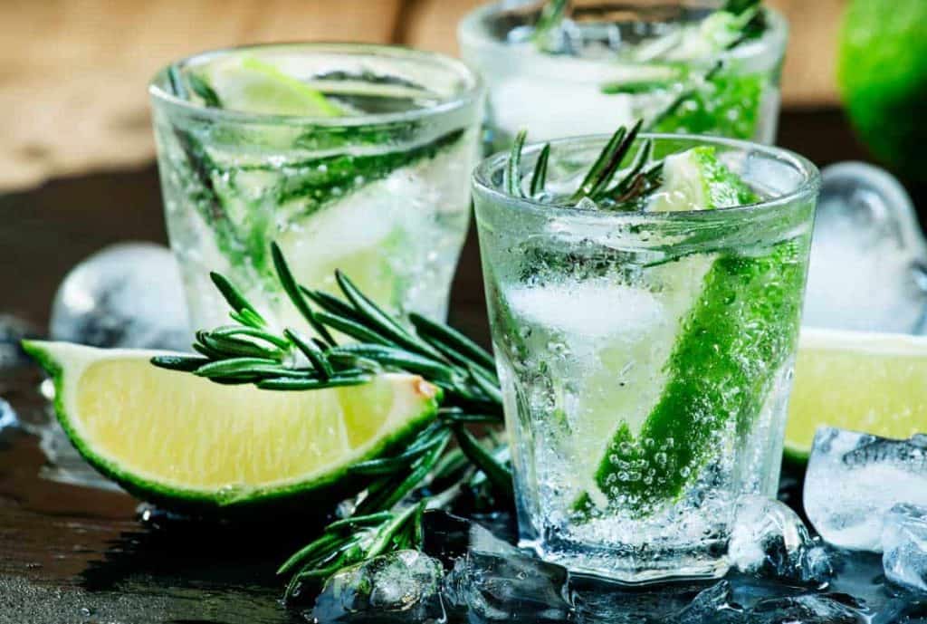 Drei Gläser Sprudelwasser mit Eis, Limettenschnitzen und Rosmarinzweigen. Im Vordergrund liegen zusätzliche Limettenscheiben und schmelzende Eiswürfel verstreut auf einer nassen Oberfläche.