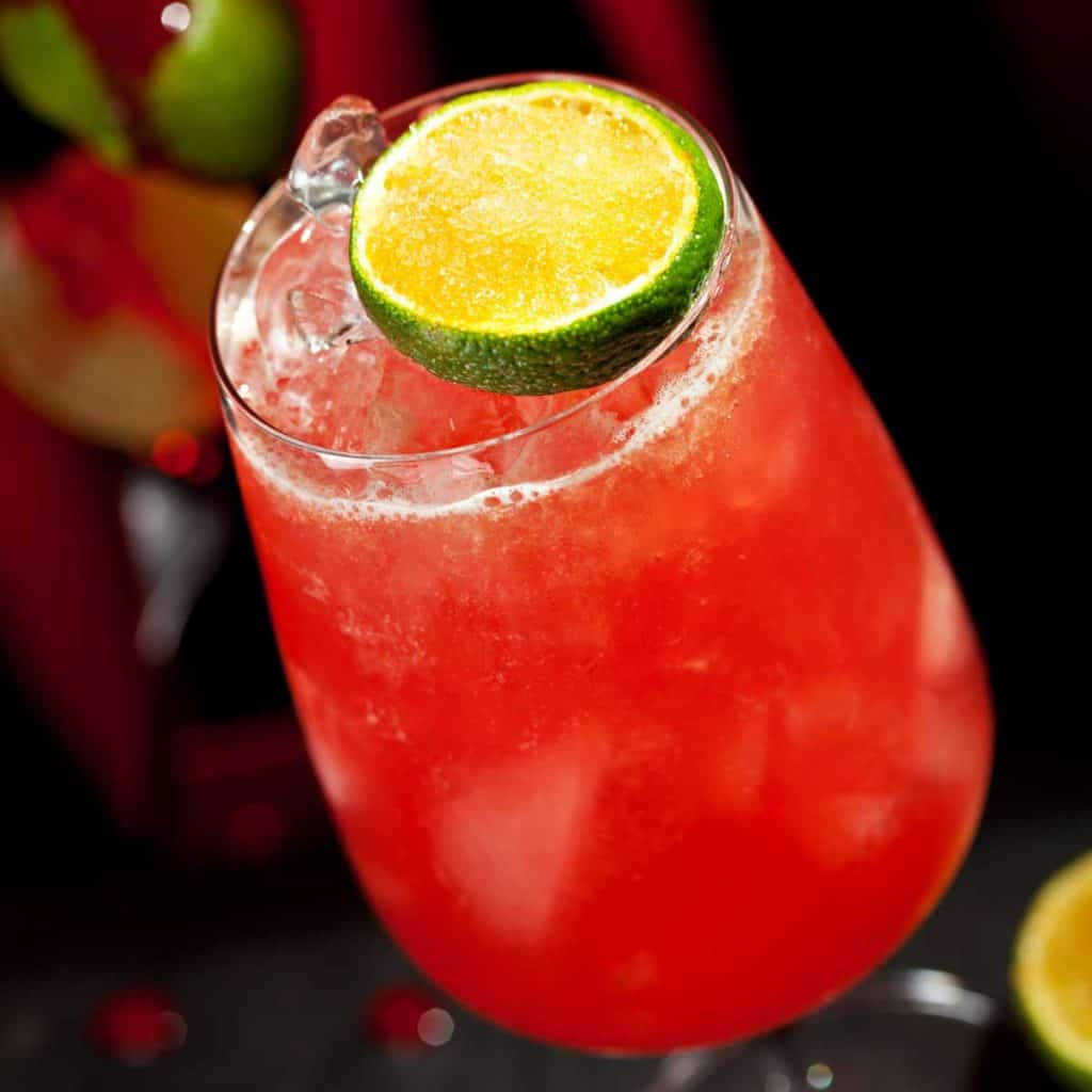Eine Nahaufnahme eines mit rotem Sirup verzierten Cocktails in einem Stielglas, garniert mit einer Limettenscheibe. Der Drink ist von einem dunklen Hintergrund umgeben, in dem andere Getränke verschwommen zu erkennen sind.