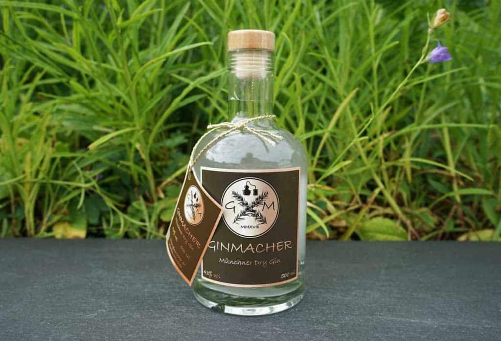 Eine Flasche „Ginmacher Munich Dry Gin“ mit braunem Etikett und einem Anhänger, platziert auf einer dunklen Oberfläche vor dem Hintergrund grüner Pflanzen.