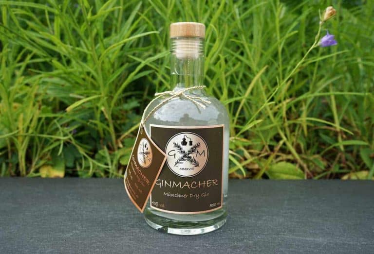 Eine Flasche „Ginmacher Munich Dry Gin“ mit braunem Etikett und einem Anhänger, platziert auf einer dunklen Oberfläche vor dem Hintergrund grüner Pflanzen.