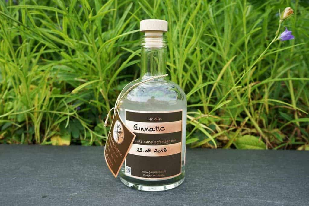 Auf einer dunklen Fläche steht eine durchsichtige Glasflasche mit der Aufschrift „Ginnatic“, einem Korken und einem braunen Etikett; im Hintergrund sind grünes Gras und violette Blumen zu sehen.
