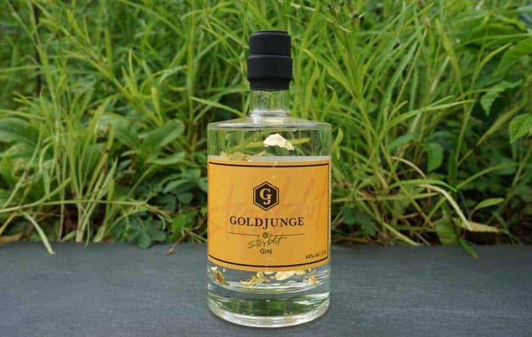 Auf einer Steinfläche steht eine Flasche Goldjunge-Gin mit gelbem Etikett und schwarzem Verschluss, im Hintergrund ist grünes Gras zu sehen. In der klaren Flüssigkeit in der Flasche sind Goldflocken zu erkennen.