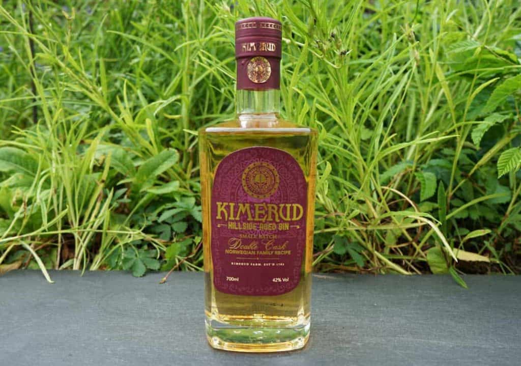 Eine Flasche „Kimerud Hillside Aged Gin“ mit einem violett-goldenen Etikett steht auf einer dunklen Fläche, umgeben von grünem Gras und Pflanzen.