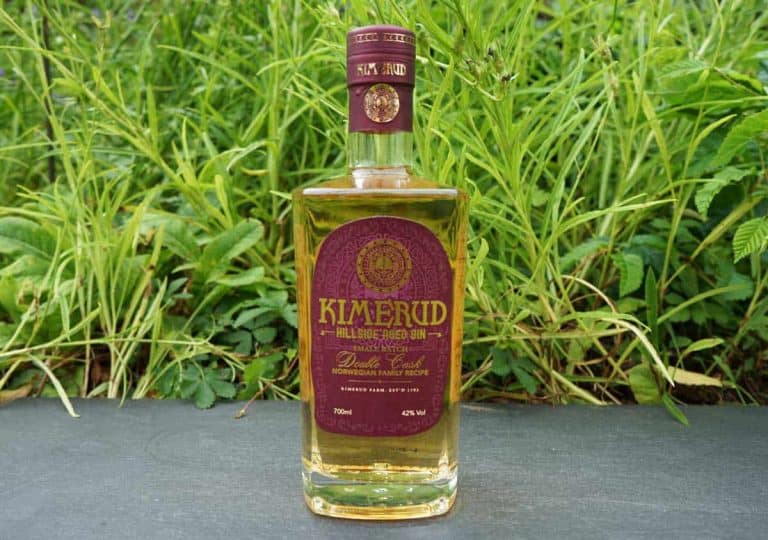 Eine Flasche „Kimerud Hillside Aged Gin“ mit einem violett-goldenen Etikett steht auf einer dunklen Fläche, umgeben von grünem Gras und Pflanzen.