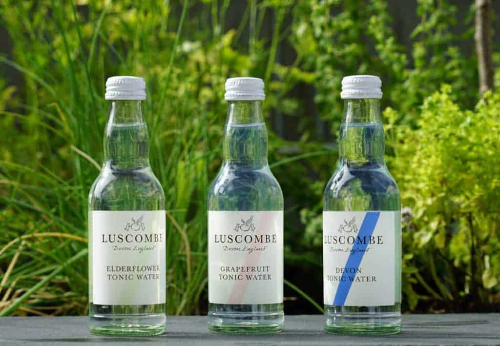 Drei durchsichtige Flaschen Luscombe-Tonic-Water – in den Geschmacksrichtungen Holunderblüte, Grapefruit und Devon – stehen im Freien auf einer dunklen Fläche, im Hintergrund sind grüne Pflanzen schemenhaft zu erkennen.