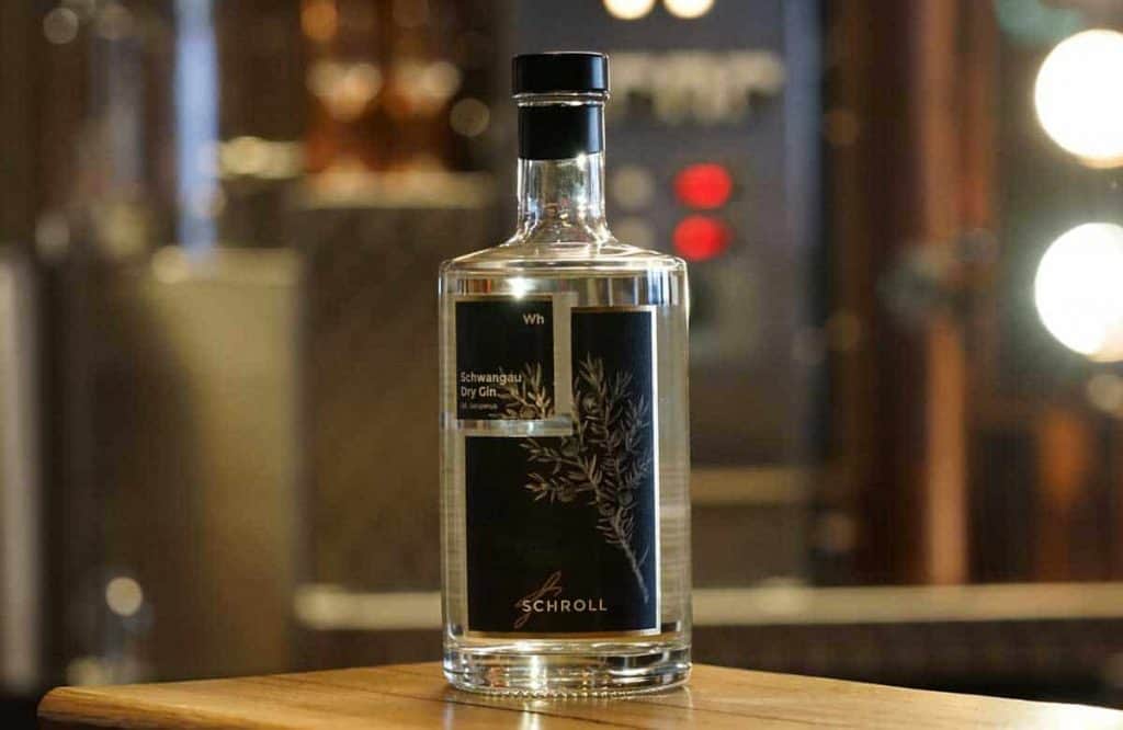 Auf einer Holzfläche steht eine klare Glasflasche mit Schroll Dry Gin und einem schwarz-goldenen Etikett, im Hintergrund sind Lichter und Metallgeräte verschwommen zu sehen.