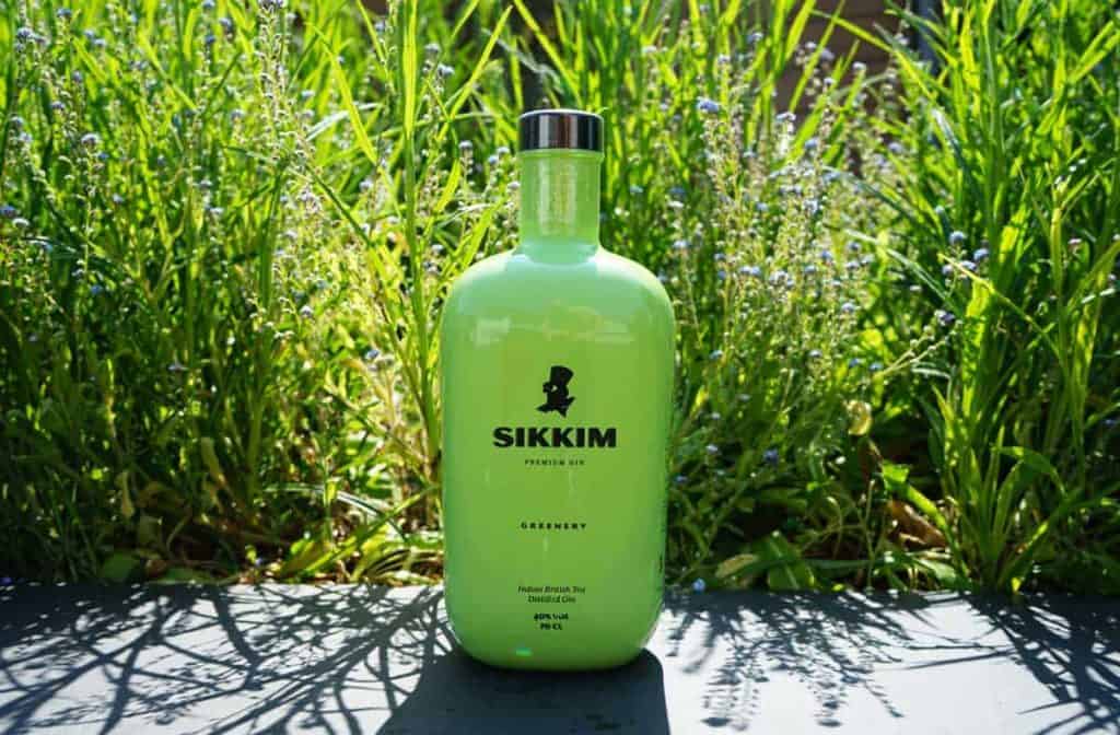 Eine grüne Flasche Sikkim-Gin steht auf einer dunklen Fläche, im Hintergrund sind hohes, grünes Gras und kleine blaue Blumen zu sehen.