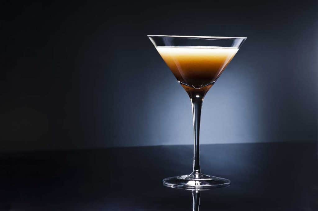 Ein Martini-Glas, gefüllt mit einem dunkelbraunen Cocktail, gekrönt von einer hellen, schaumigen Schicht, vor einem dunklen Hintergrund mit Farbverlauf und in einer glänzenden Oberfläche gespiegelt.