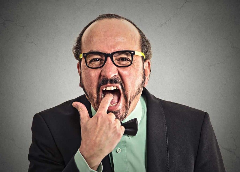 Ein Mann mittleren Alters mit Brille und Anzug zeigt mit dem Finger in seinen offenen Mund und verzieht dabei angewidert das Gesicht, als würde er so tun, als müsse er würgen, oder als wolle er starke Abneigung oder Missbilligung zum Ausdruck bringen. Grauer Hintergrund.