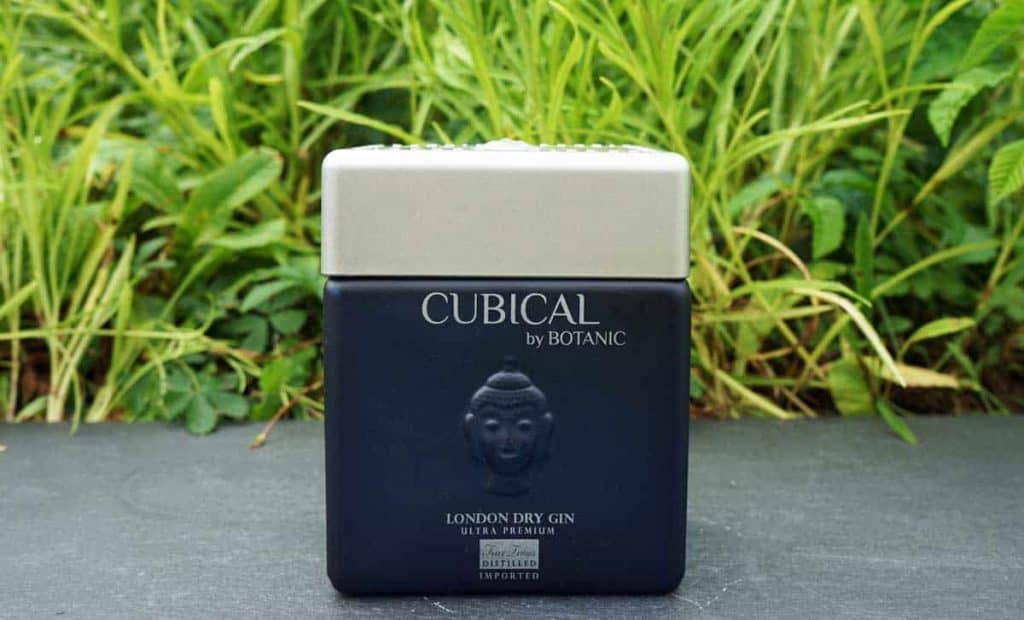 Eine quadratische, dunkelblaue Flasche „Cubical“ von Botanic London Dry Gin steht auf einer grauen Fläche, im Hintergrund sind grüne Pflanzen zu sehen. Die Flasche ist mit einem Buddha-Gesicht verziert und hat einen silbernen Verschluss.