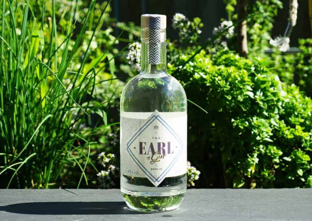 Eine Flasche „The Earl Gin“ steht im Freien auf einer dunklen Oberfläche, umgeben von üppig grünen Pflanzen und im Sonnenlicht.
