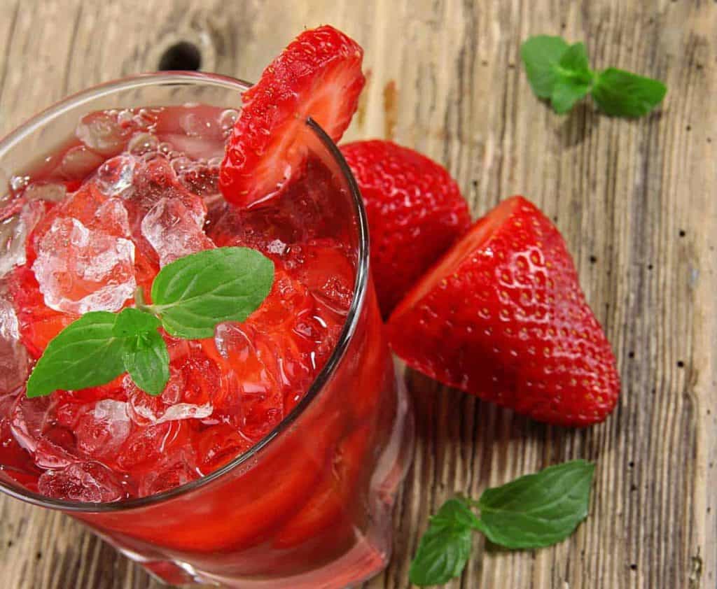 Ein Glas Erdbeergetränk mit Eis, garniert mit Minze und einer Erdbeerscheibe, steht auf einer rustikalen Holzunterlage neben ganzen Erdbeeren und Minzblättern.