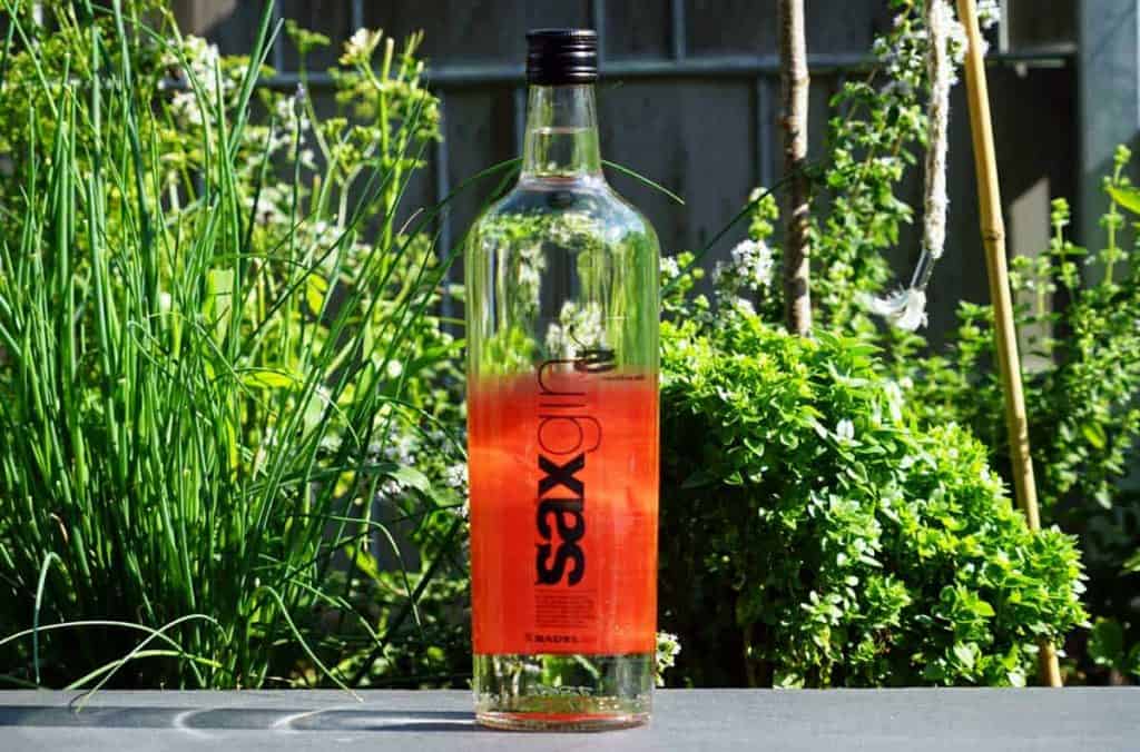 Eine durchsichtige Flasche mit der Aufschrift „Sex on“ und einem schwarzen Verschluss, in deren Boden sich eine rote Flüssigkeit befindet, steht im Freien auf einer Fläche, im Hintergrund sind grüne Pflanzen zu sehen.