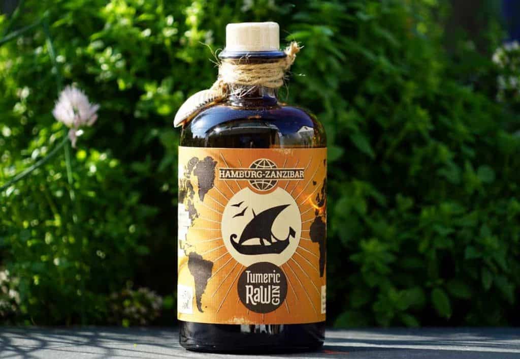 Auf einem Tisch im Freien steht eine dunkle Glasflasche mit der Aufschrift „Hamburg-Zanzibar Turmeric Raw Gin“ und einem stilisierten Segelboot-Logo, im Hintergrund sind üppig grüne Pflanzen zu erkennen.