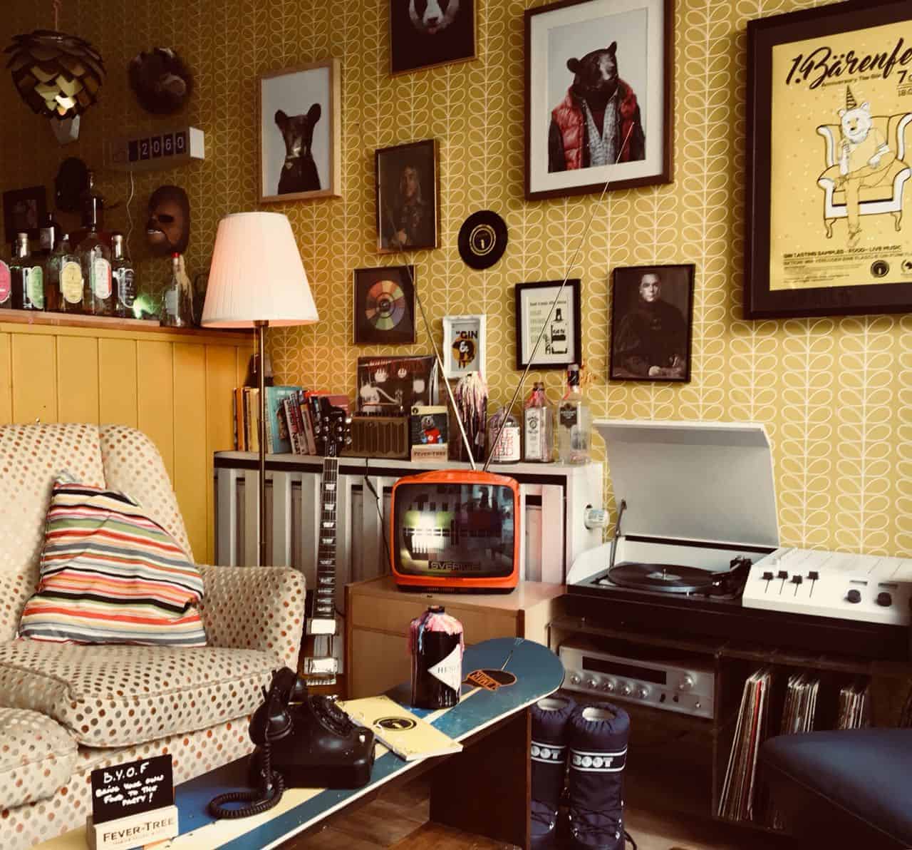 Ein gemütliches Wohnzimmer im Retro-Stil mit gemusterter Tapete, Vintage-Dekoration, einem gestreiften Sessel, einem Plattenspieler, einem kleinen Fernseher, Regalen mit Flaschen, gerahmten Tierporträts sowie verschiedenen Büchern und Schallplatten.