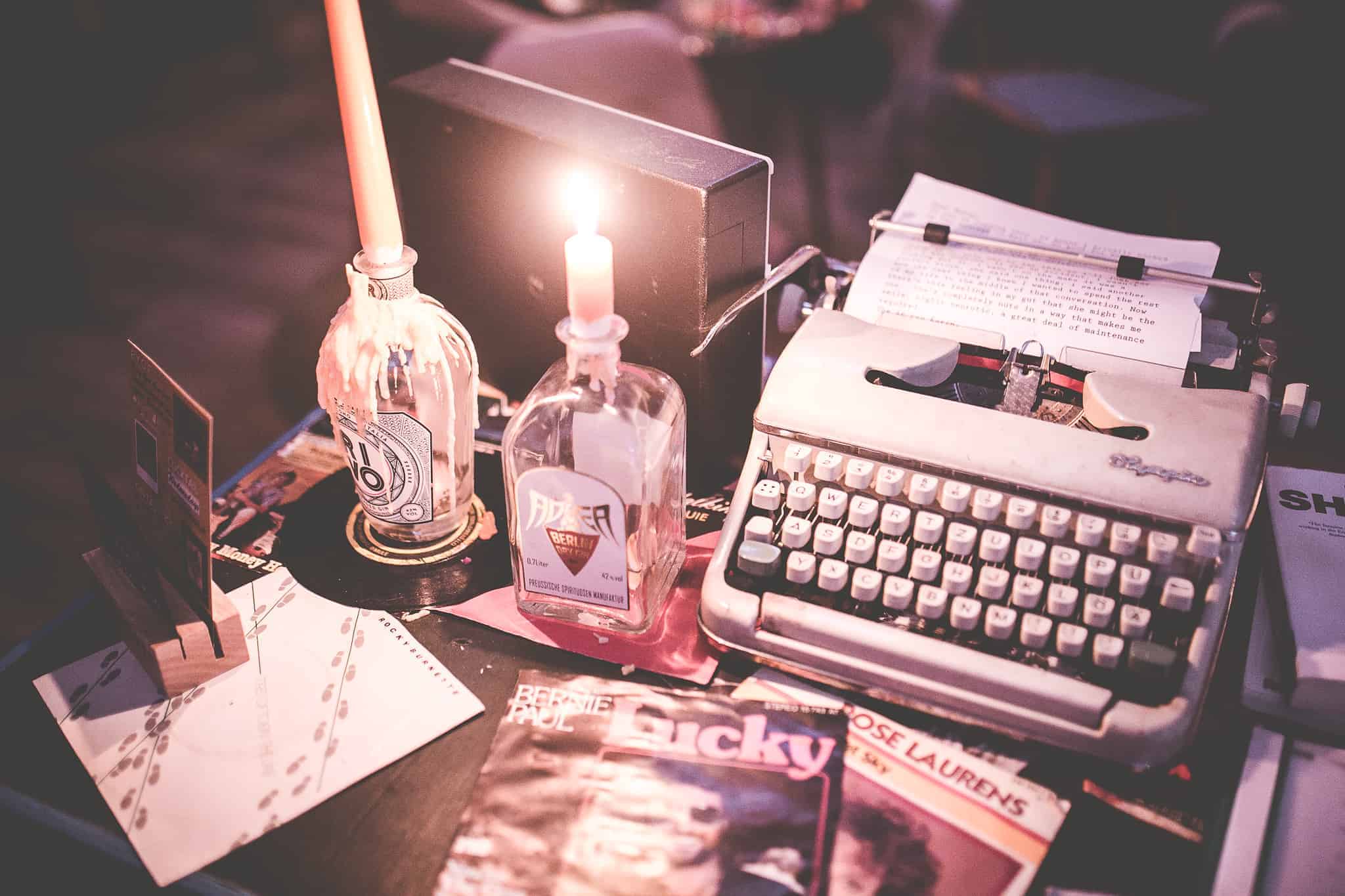 Eine alte Schreibmaschine, eine brennende Kerze in einer Flasche und verstreute Zeitschriften liegen auf einem dunklen Tisch und schaffen mit ihrem warmen, sanften Licht eine nostalgische, gemütliche Atmosphäre.