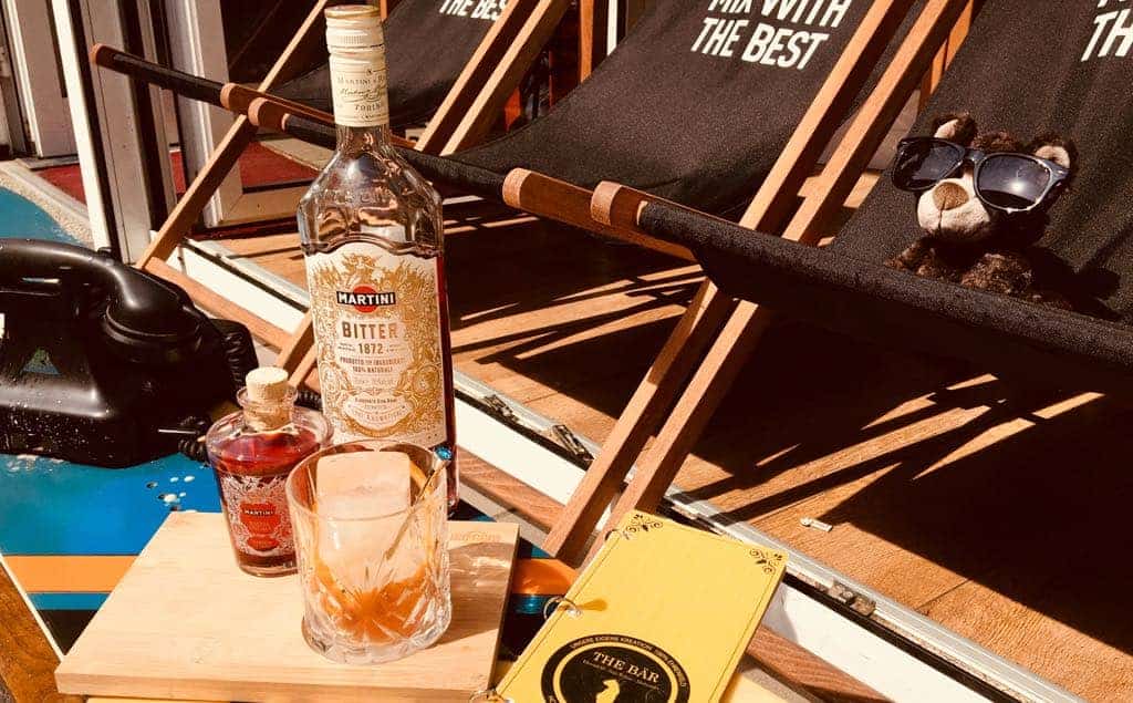 Auf einem kleinen Tisch neben schwarzen Liegestühlen stehen ein Cocktail, Schnapsflaschen und eine gelbe Speisekarte. Auf einem der Stühle liegt ein Teddybär mit Sonnenbrille. Sonnenlicht taucht die entspannte Szene am Pool in ein helles Licht.