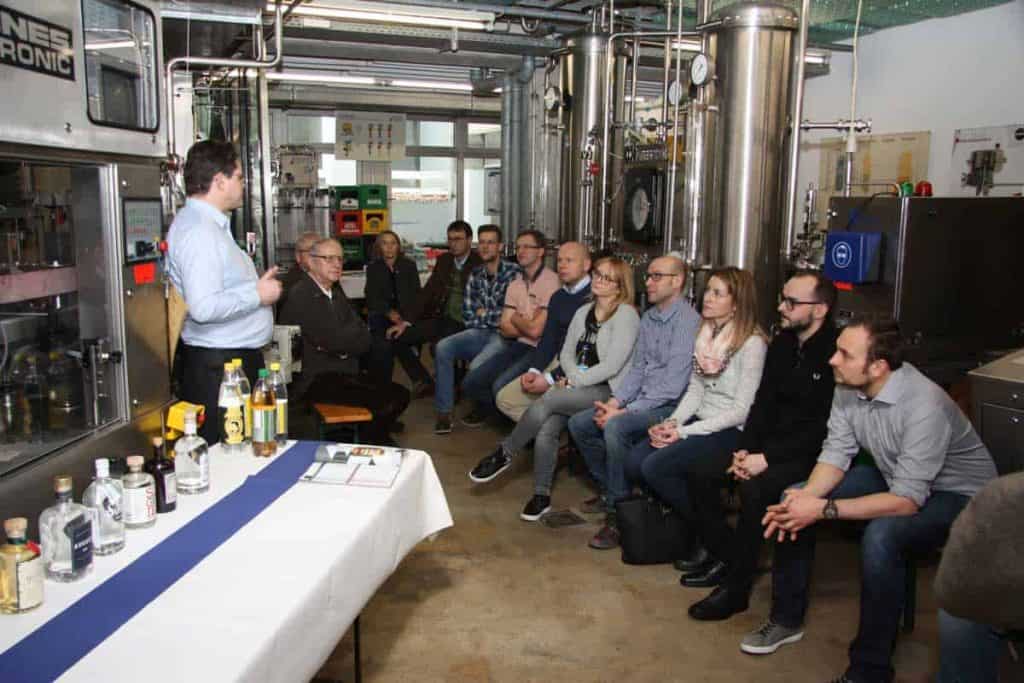 In einem Industrieraum mit großen Metalltanks sitzt eine Gruppe von Menschen und hört einem Mann zu; auf einem Tisch im Vordergrund stehen verschiedene Flaschen.