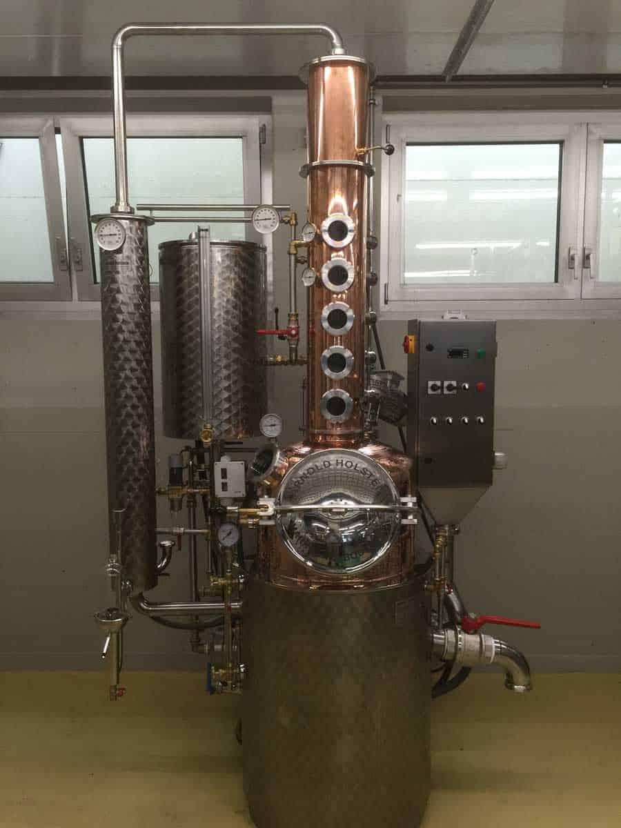 Ein Destillierkolben aus Kupfer und Edelstahl mit Messgeräten, Rohren und einem Bedienfeld, aufgestellt in einem sauberen Industrieraum mit Fenstern im Hintergrund.