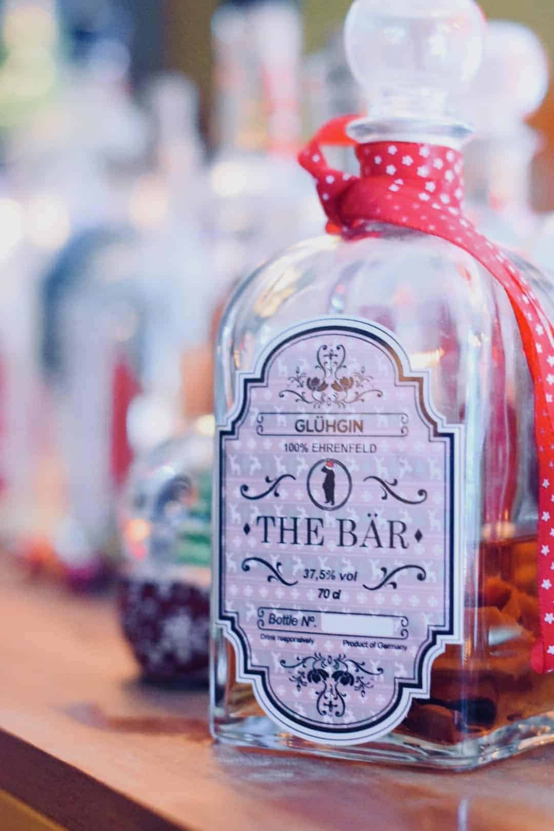 Auf einer Holzfläche steht eine Glasflasche mit der Aufschrift „THE BAR“, um deren Hals ein rotes Band mit weißen Sternen gebunden ist; im Hintergrund sind verschwommene Flaschen zu sehen.