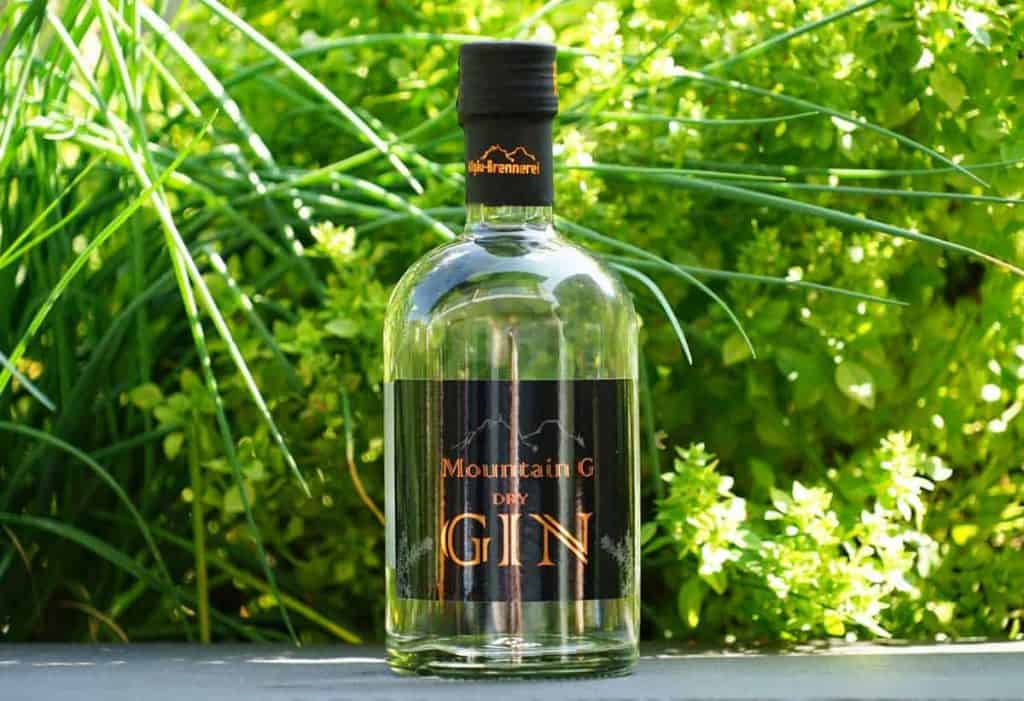 Auf einer grauen Fläche steht eine klare Glasflasche mit „Mountain G Dry Gin“, im Hintergrund ist leuchtend grünes Laub zu sehen.