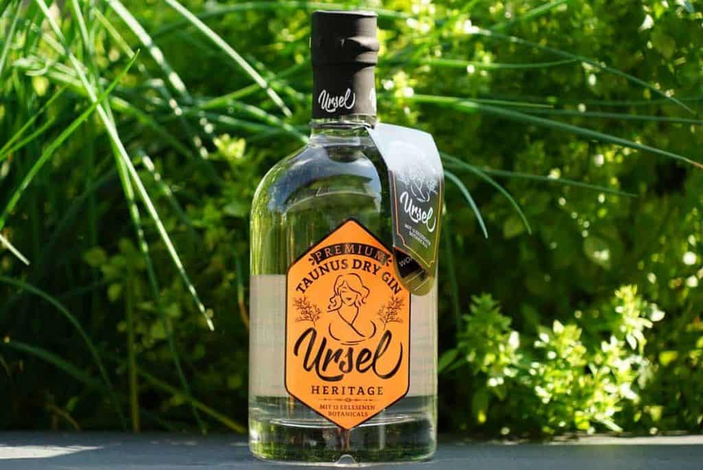 Eine klare Glasflasche „Ursel Heritage Taunus Dry Gin“ mit orangefarbenem Etikett steht auf einer dunklen Fläche, im Hintergrund umgeben von üppigen grünen Pflanzen.