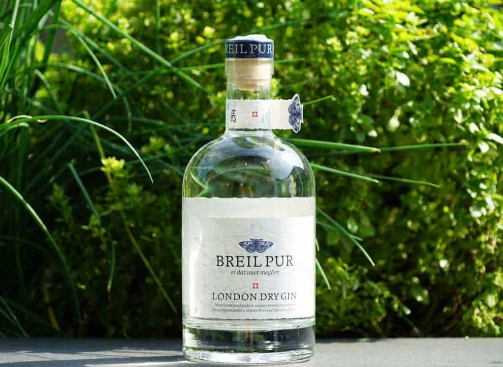 Eine Flasche Breil Pur London Dry Gin steht im Freien auf einer Fläche, umgeben von üppig grünen Pflanzen.