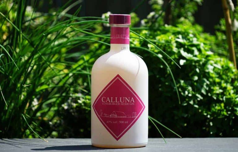 Eine weiße Flasche „Calluna Lüneburger Heide Gin“ mit einem rosa Etikett steht im Freien auf einer dunklen Fläche, im Hintergrund von grünen Pflanzen umgeben.