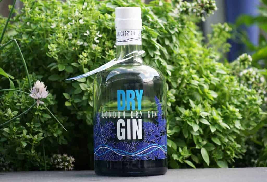 Eine durchsichtige Flasche London Dry Gin mit blau-weißem Etikett steht im Freien vor üppig grünen Kräutern und Pflanzen.