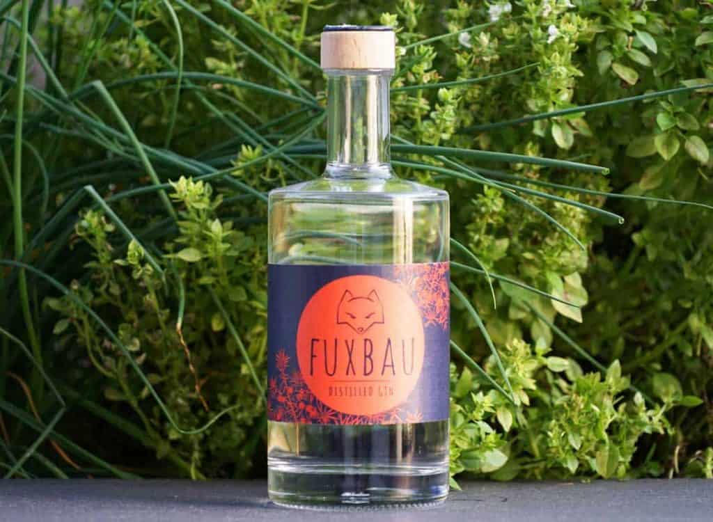 Vor grünen Pflanzen steht eine klare Glasflasche mit Fuxbau-Gin und einem Holzkorken. Das Etikett ist orange und schwarz und zeigt die Abbildung eines Fuchses sowie den Schriftzug „Fuxbau Distilled Gin“.