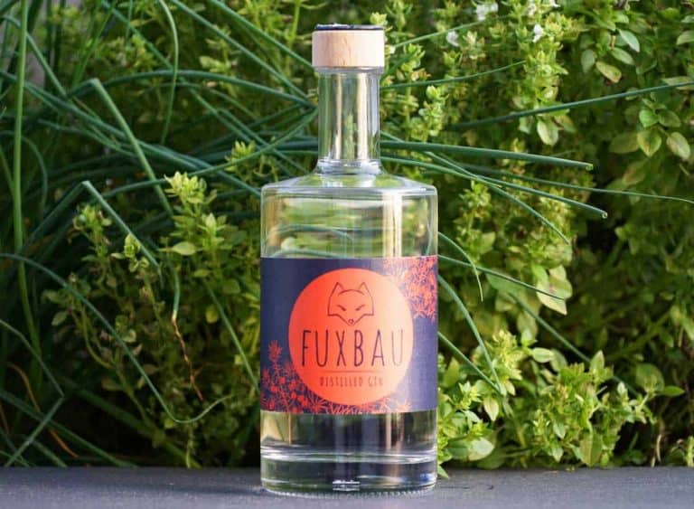 Vor grünen Pflanzen steht eine klare Glasflasche mit Fuxbau-Gin und einem Holzkorken. Das Etikett ist orange und schwarz und zeigt die Abbildung eines Fuchses sowie den Schriftzug „Fuxbau Distilled Gin“.