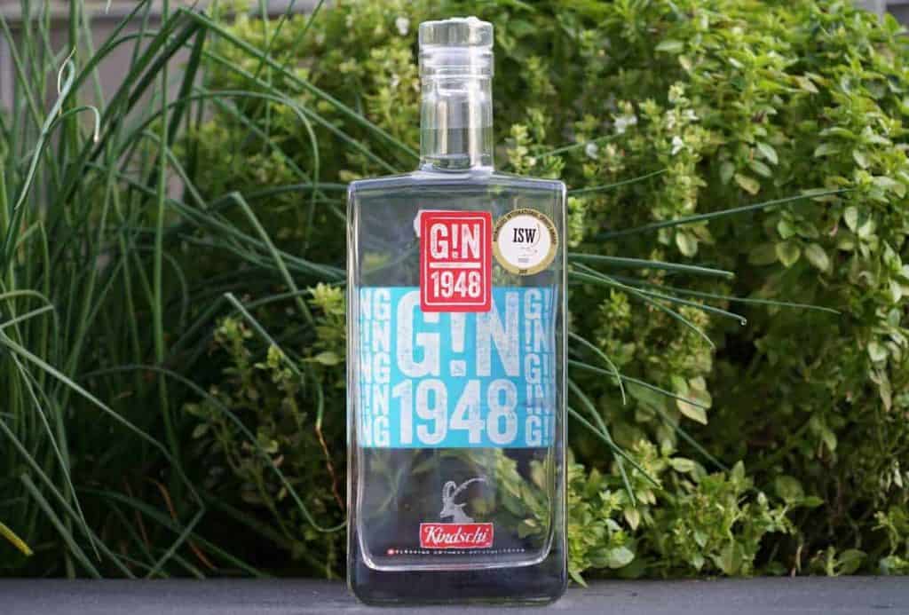 Vor üppig grünen Kräutern steht eine rechteckige Flasche „GIN 1948“ mit blauen und roten Etiketten. Das Etikett ist mit einer auffälligen Schrift versehen und weist oben einen kleinen goldenen Auszeichnungsaufkleber auf.