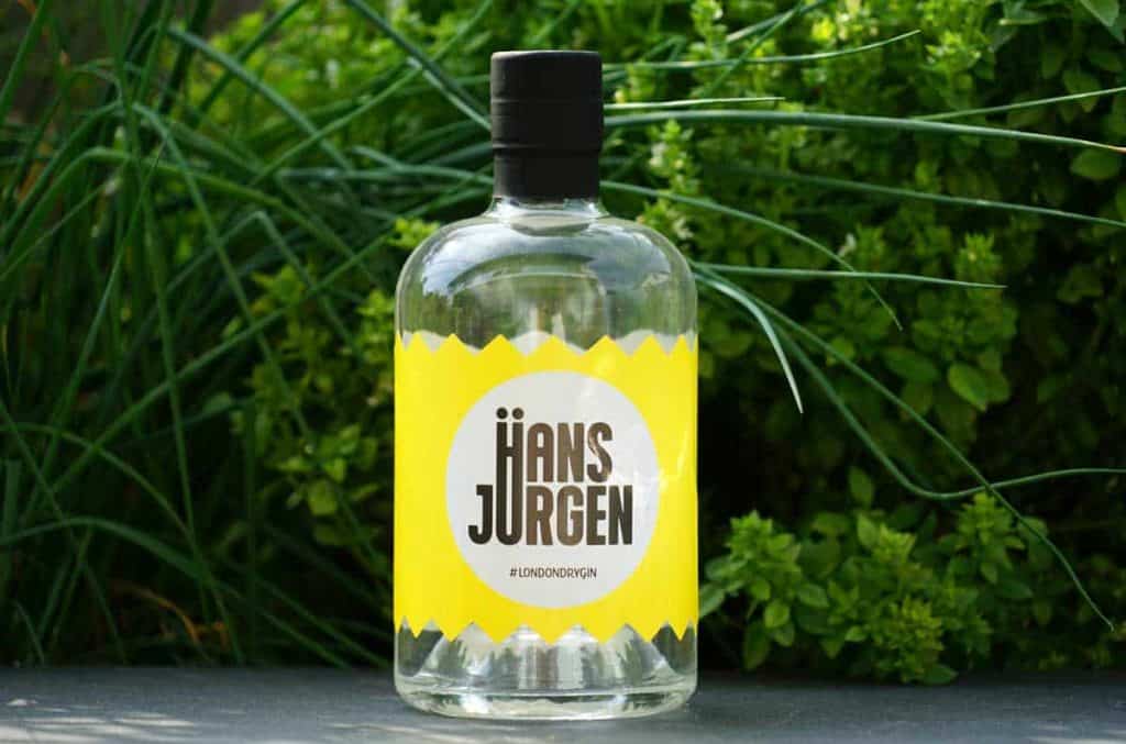 Auf einer dunklen Fläche steht eine durchsichtige Glasflasche mit der Aufschrift „HäNS JüRGEN“, im Hintergrund sind grüne Pflanzen zu sehen. Das Etikett ist gelb und mit fetter schwarzer Schrift bedruckt.
