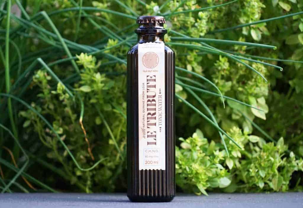 Eine Flasche „Le Tribute“-Tonic Water steht im Freien aufrecht, im Hintergrund sind grüne Blattpflanzen zu sehen.