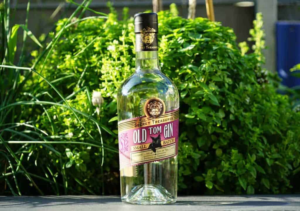 Eine Flasche „Secret Treasures Old Tom Gin“ steht im Freien auf einer ebenen Fläche, im Hintergrund sind üppig grüne Pflanzen und Laub zu sehen.