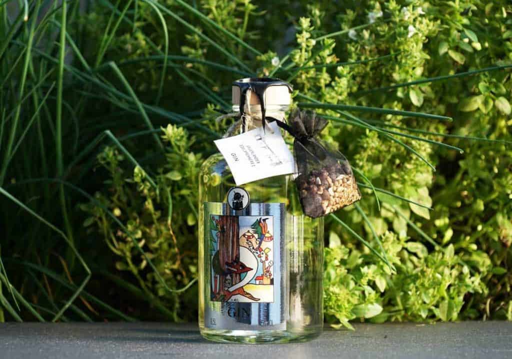 Eine Flasche Gin mit einem bunten Etikett und einem kleinen Säckchen mit Botanicals, die vor üppig grünen Kräutern steht.